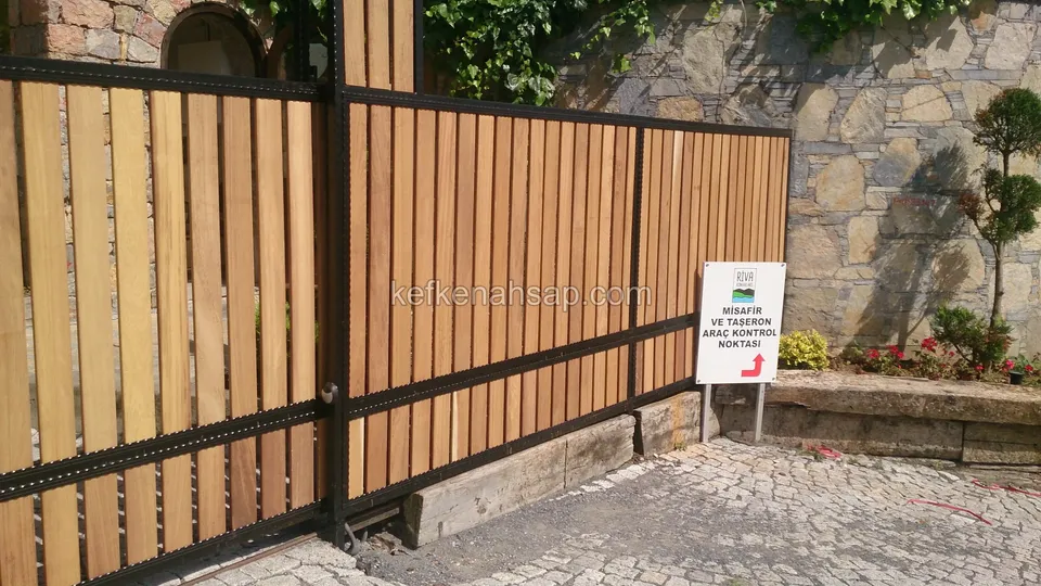 Taş duvarın önünde, küp taş yola paralel uzanan siyah çerçeveli dikey çıtalı çit; yanda küçük bir tabela, yerde kalas