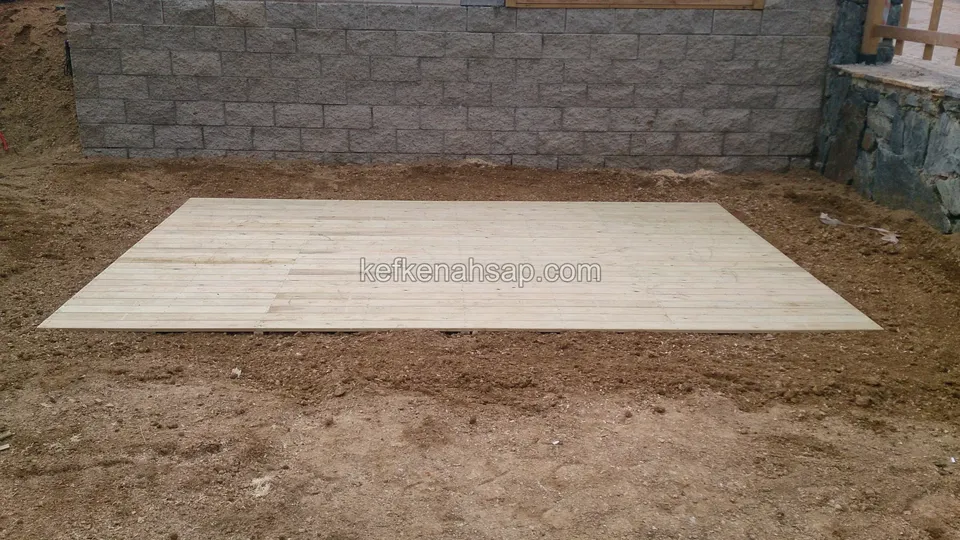Taş duvarın önünde, çıplak toprak zemine serilmiş küçük açık renkli deck; çevre henüz düzenlenmemiş