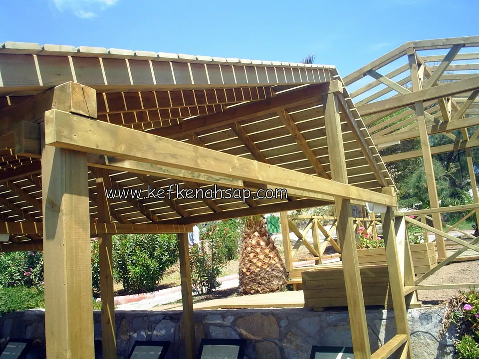 Taş duvar ve palmiyenin önünde tahta çatılı ahşap pergola, çevrede çiçekli bitkiler ve altta şezlonglar
