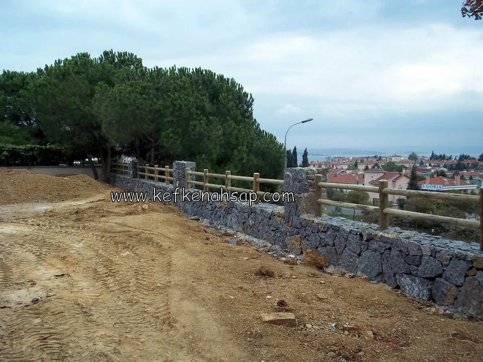 Yamaç kenarındaki taş duvarın üstünde uzanan, taş sütunlarla ayrılmış yatay kirişli ahşap korkuluk, arkada şehir ve deniz