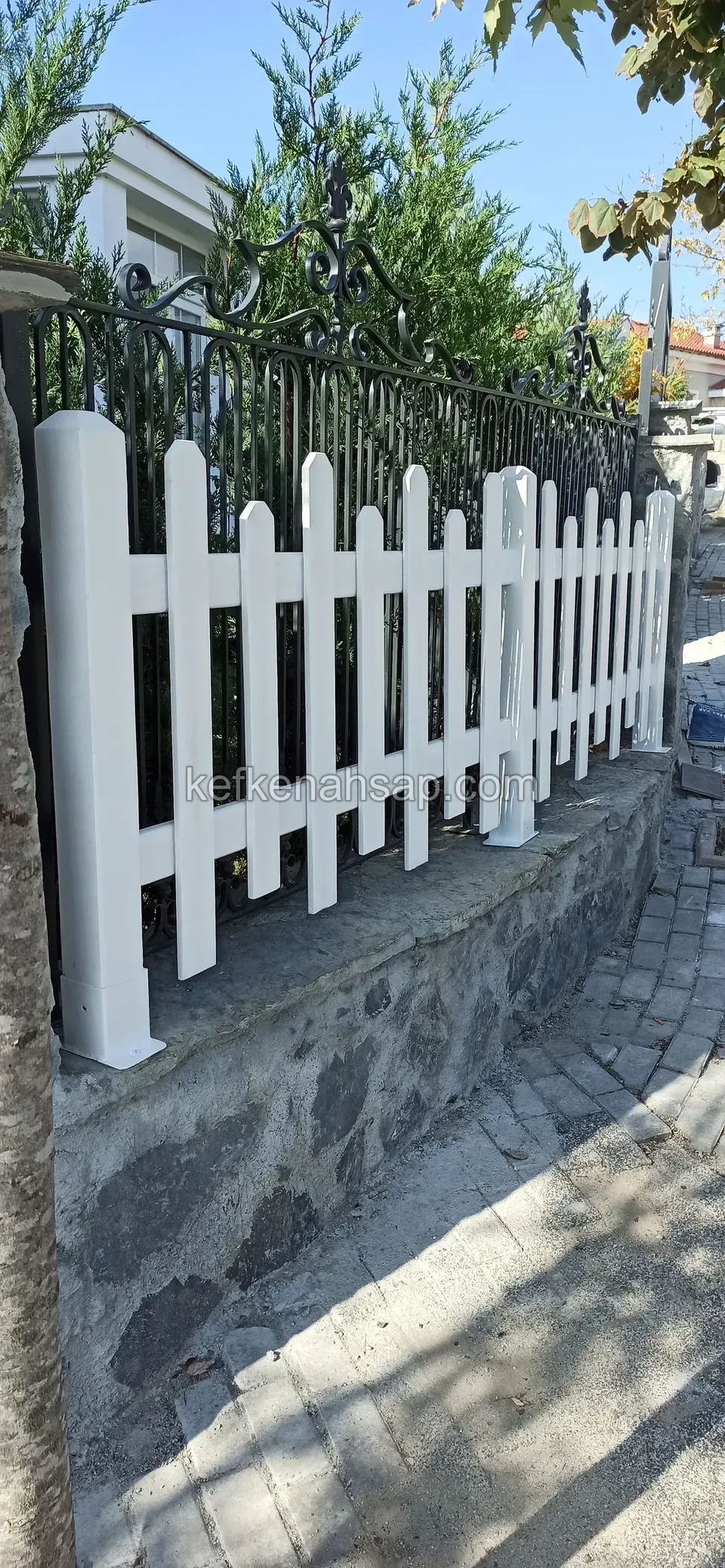Taş duvarın üstünde beyaz boyalı ahşap çıtalı çit, arkasında siyah demir parmaklık ve yeşillik