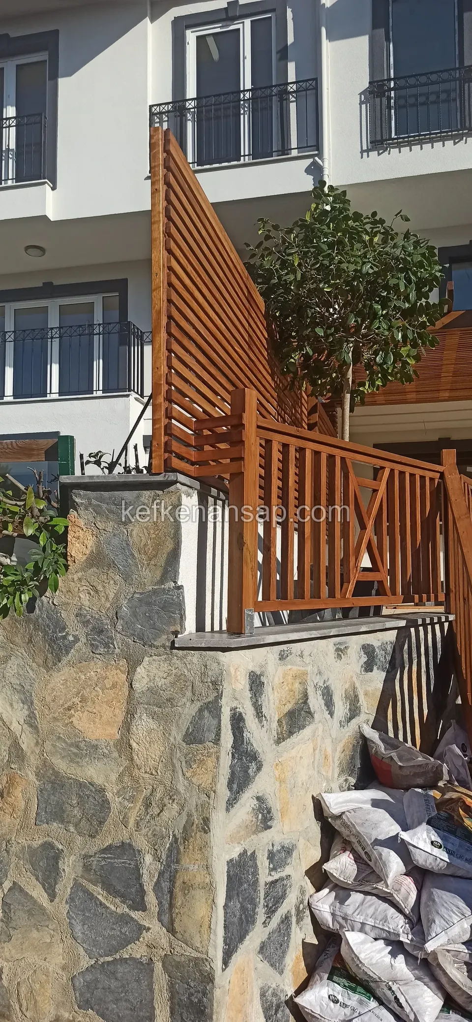 Taş duvarın üstünde turuncu tonlu dikey çıtalı korkuluk ve yanında yatay çıtalı yüksek paravan