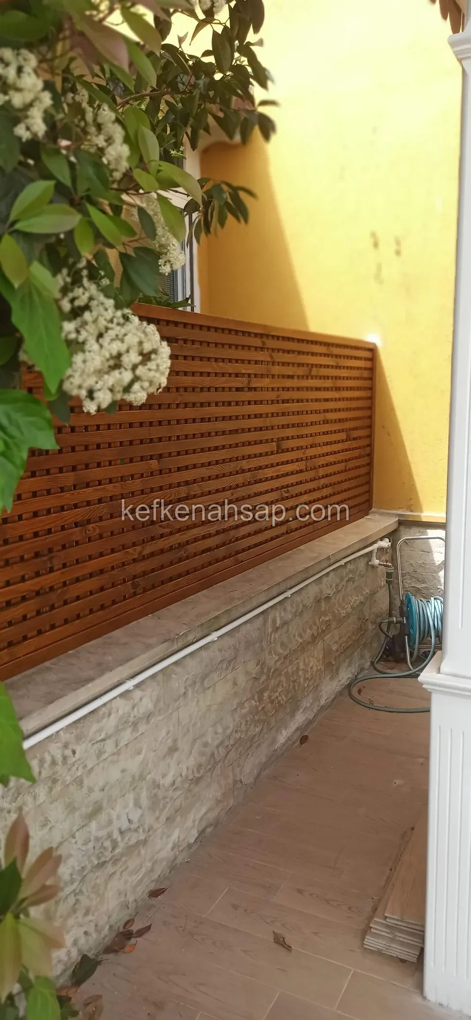 Taş kaplı alçak duvarın üstünde uzanan sık çıtalı turuncu ahşap panel; arkada sarı cephe, önde çiçekli ağaç dalları