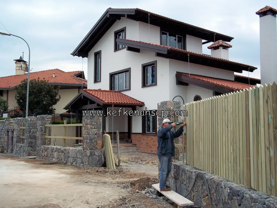 Taş duvarın üzerine sivri uçlu açık renk dikey çıtalı çit çakılıyor; çalışan usta ve arkada kiremit çatılı iki katlı villa