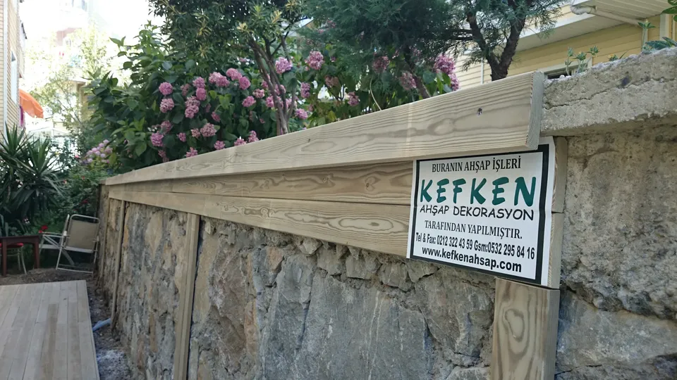 Taş duvarın üzerine oturtulmuş açık renk ahşap tahta ve üzerindeki yazılı pano