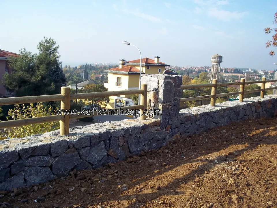 Yakından görünen taş duvar ve üstündeki yatay kirişli ahşap çit, arkada su kulesi ve evler