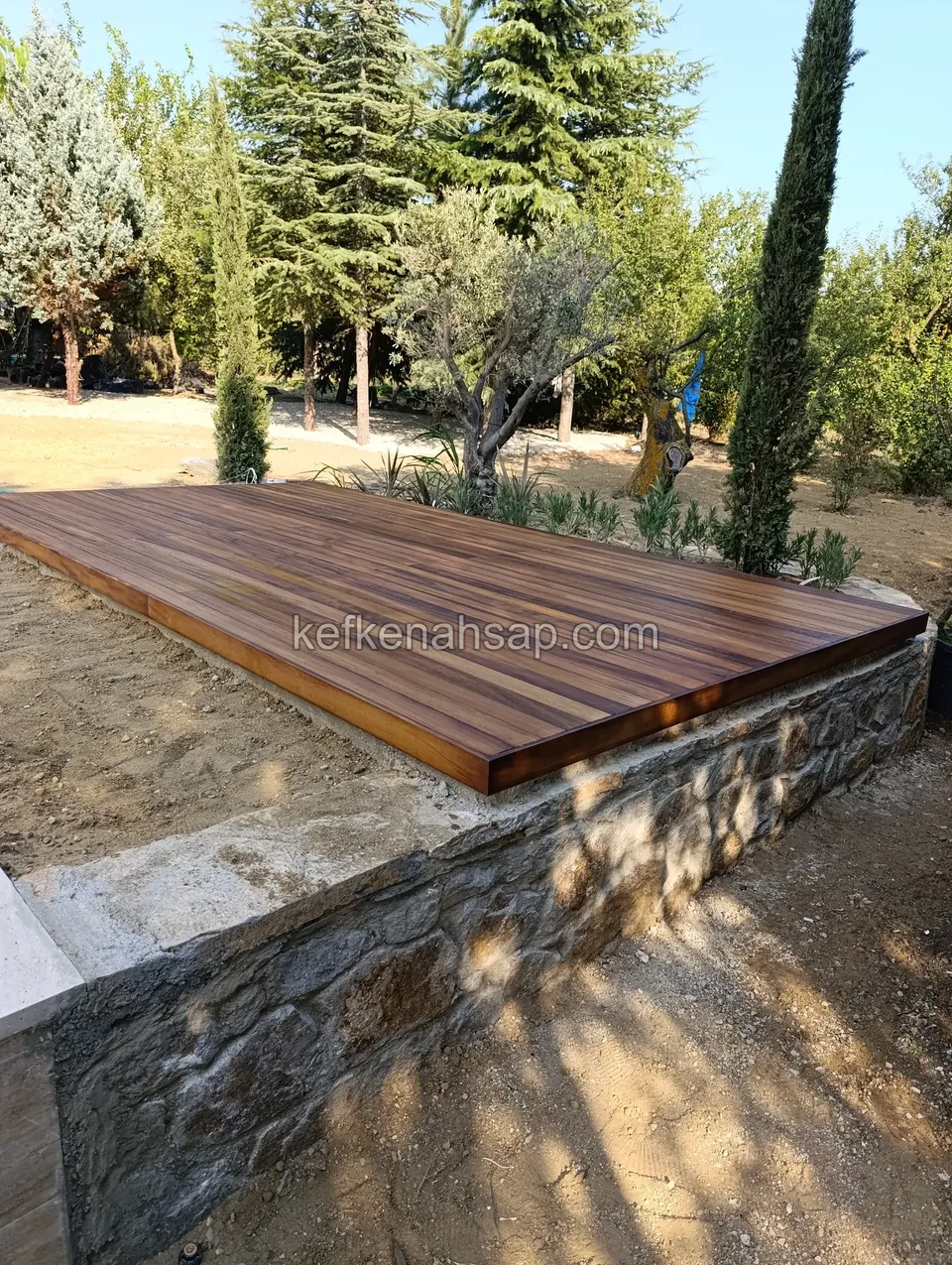 Dark red-brown deck resting on a low natural-stone wall, with pine trees, olives and cypresses behind