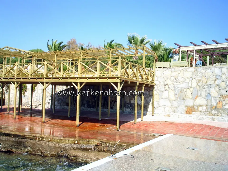 Taş duvarın yanında, uzun ayaklar üzerinde yükselen X korkuluklu ve pergola çatılı deck, altta kırmızı zemin ve su kenarı