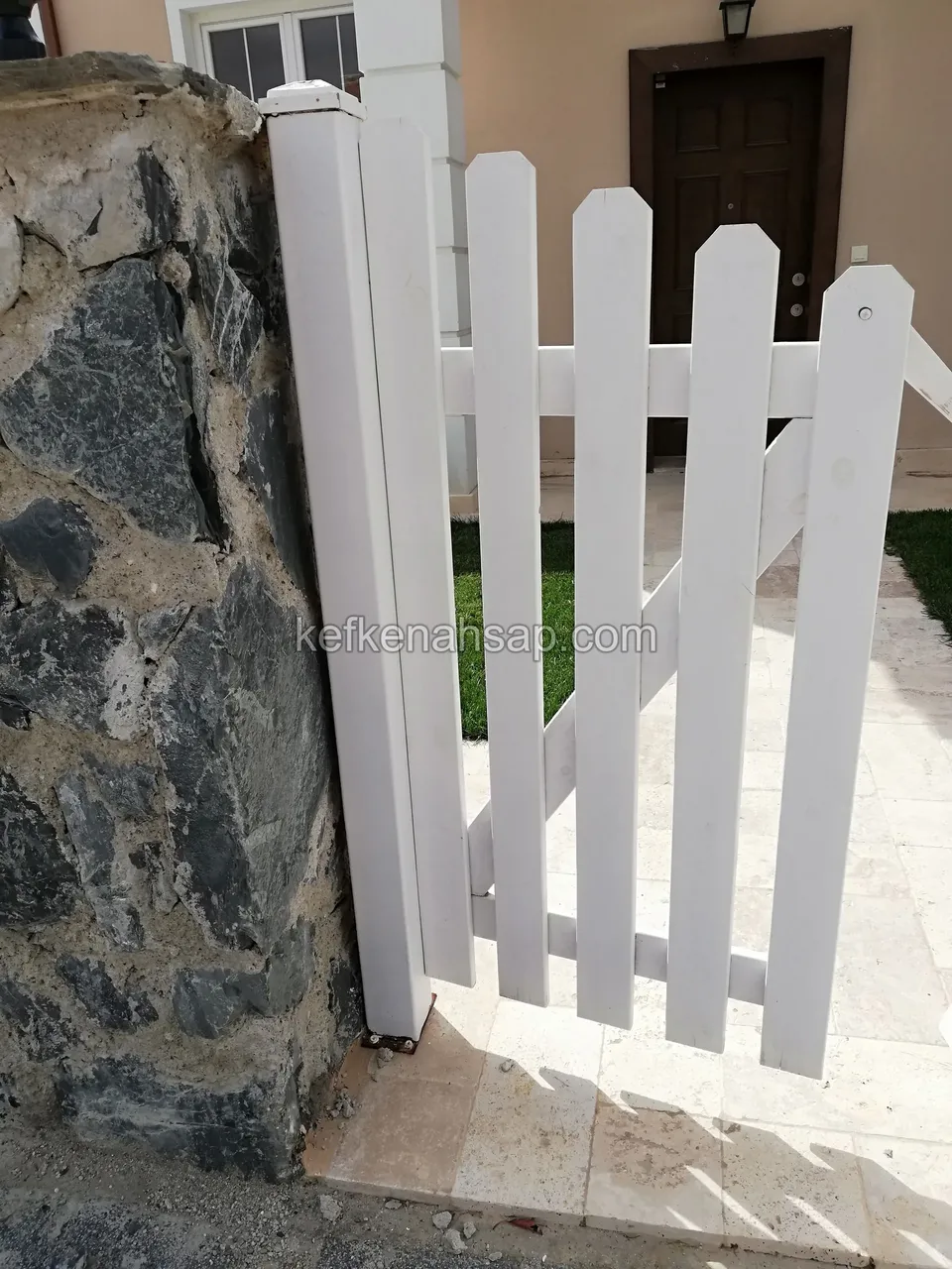 Koyu taş duvarın yanında, kalın beyaz direğe bağlı ucu kavisli çıtalı beyaz bahçe kapısı, arkada ev girişi