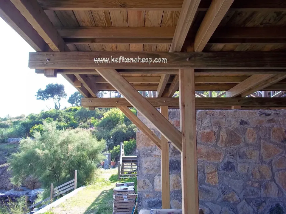 Taş duvarın yanında pergola kirişi, çapraz payanda ve tahta kaplı çatı alt kısmı, arkada çalılık ve kıyı
