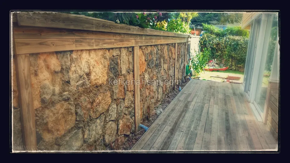 Light grey-toned deck laid in the narrow space between a stone wall and a glass door, with a timber rail on top of the wall