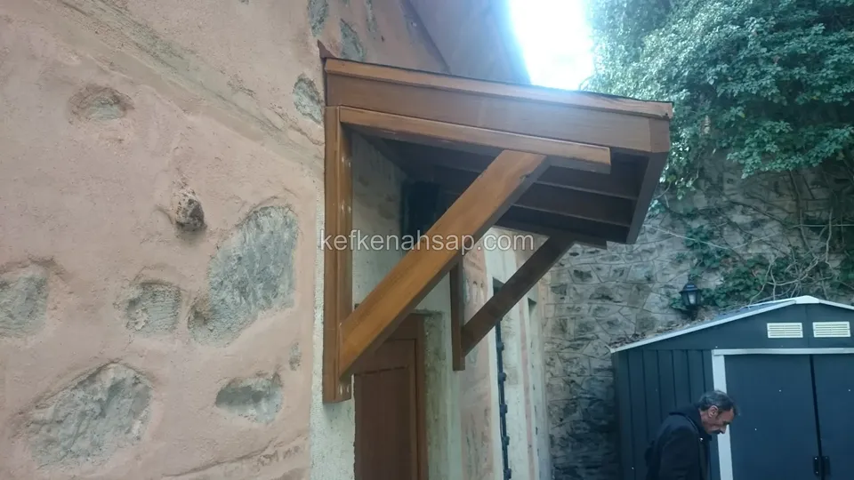 Taş örgülü duvarda bir kapının üzerinde, ahşap konsollarla taşınan eğimli ahşap saçak; yanda gri bahçe kulübesi