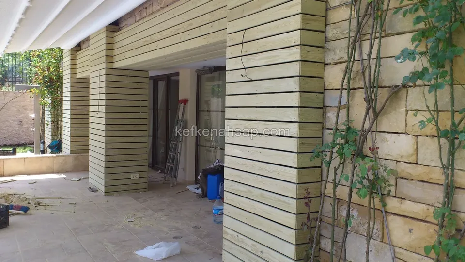 Taş duvar ve sarmaşığın yanında, yatay çıtalarla kaplanmış kalın kolonlar ve camlı kapılı teras girişi, altta işçi aletleri