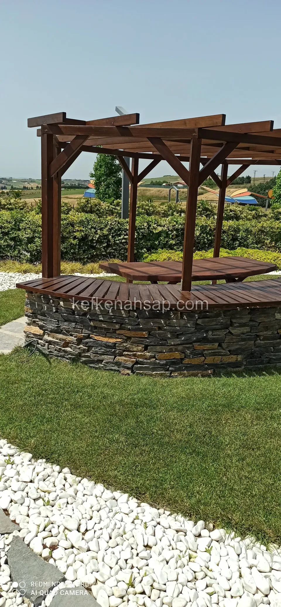 Bahçede taş duvarlı yuvarlak bank üzerinde ahşap pergola, çevrede çim, beyaz çakıl ve çit