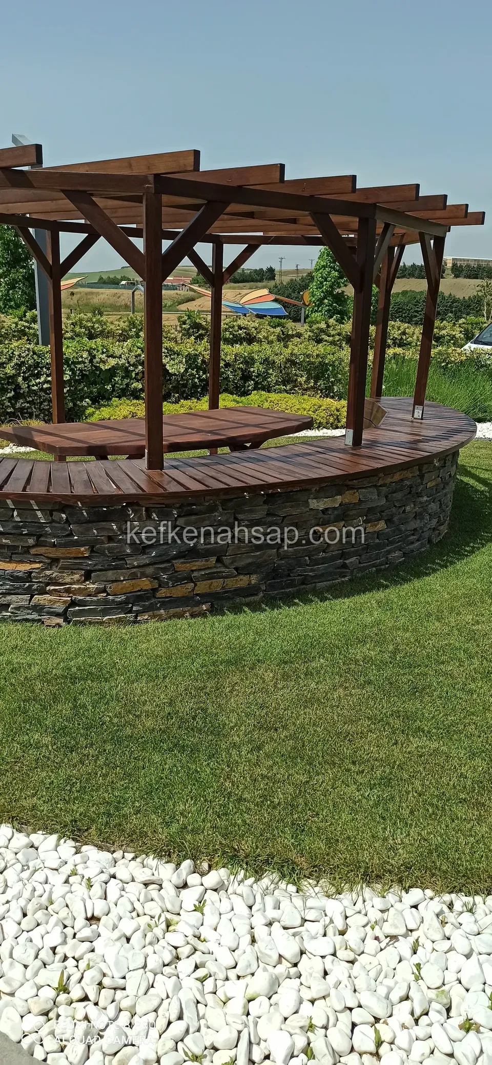 Taş örgülü kavisli ahşap bankın üstünde vernikli pergola, çimlik ve beyaz çakıl şeridi