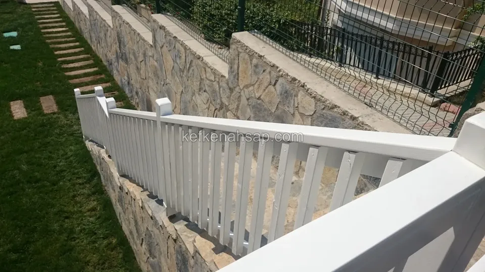 Taş merdiven boyunca uzanan beyaz boyalı çıtalı ahşap korkuluk; yanda çimlik ve taş duvar