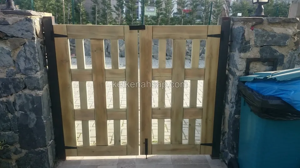 Koyu taş sütunlar arasında, emprenyeli çam görünümlü açık renk bölmeli çift kanatlı ahşap kapı; sağda mavi torbalı çöp kutusu