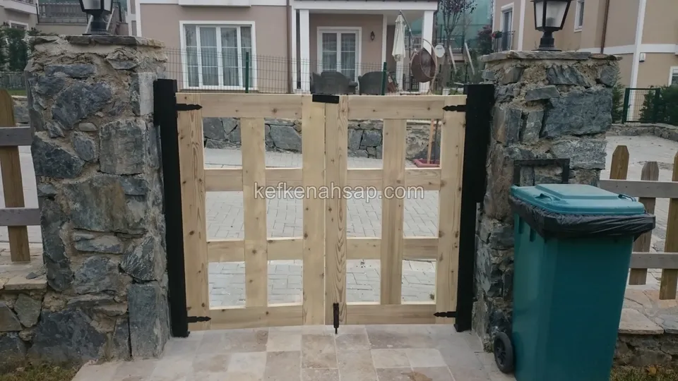 Koyu taş sütunlar arasında kapalı duran, bölmeli açık renk ahşap çift kanatlı kapı; sağda çöp konteyneri, arkada ev