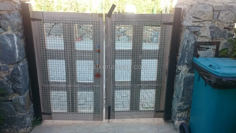 Koyu taş sütunlar arasında, kare bölmeli ve tel örgü dolgulu gri tonlu çift kanatlı ahşap kapı; sağda yeşil çöp konteyneri