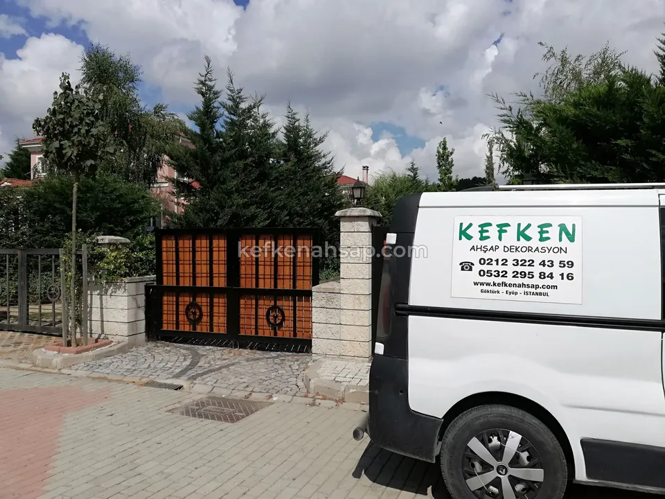 İki taş sütun arasında, turuncu kare dolgulu koyu çerçeveli çift kanatlı bahçe kapısı; önde beyaz bir araç