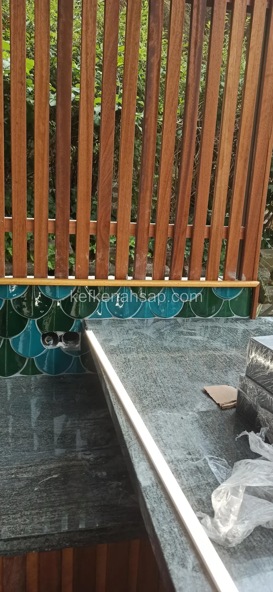 Taş tezgâhın kenarında parlak metal profil, arkada dikey ahşap çıtalar ve yeşil seramik, yanda ambalaj artığı
