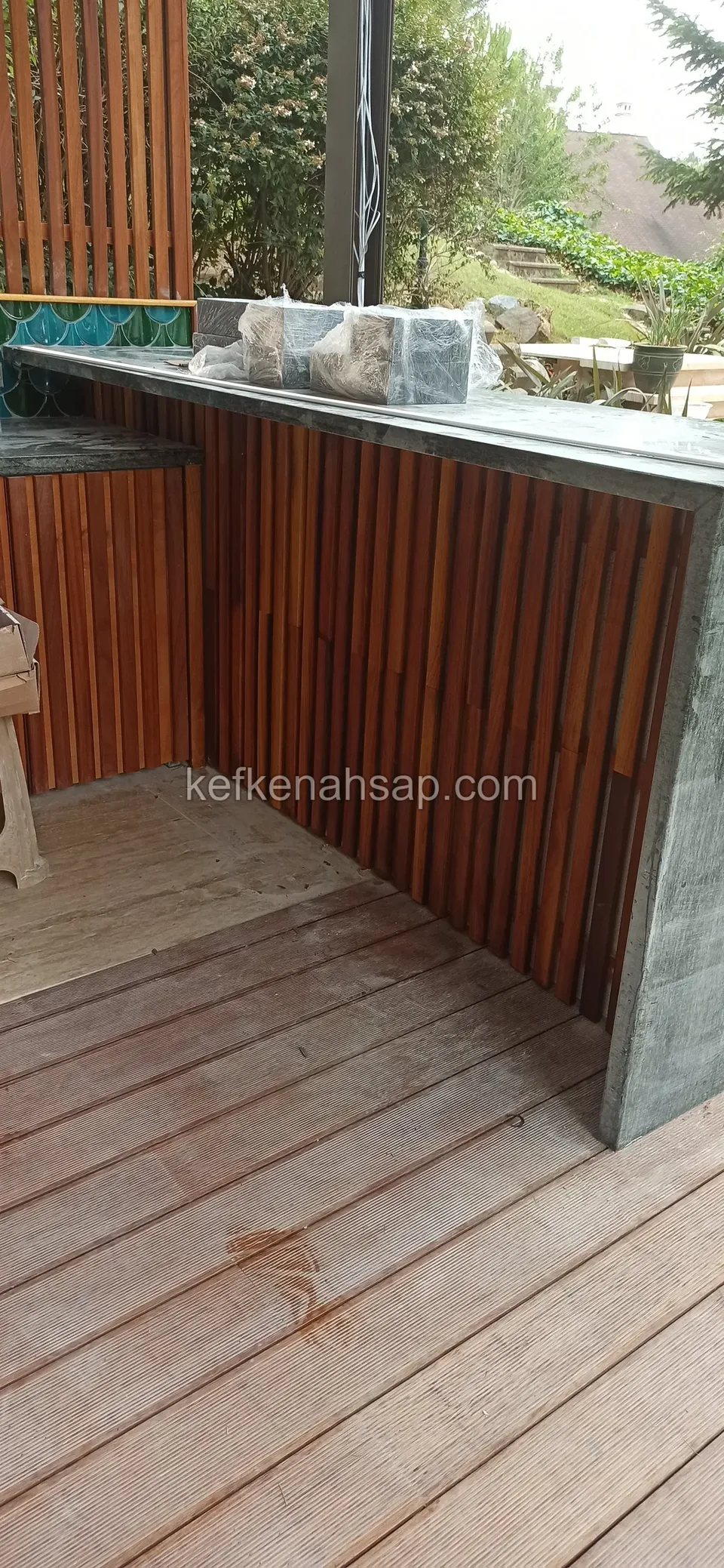 Deck zemin üzerinde, önü dikey ahşap şeritlerle kaplı taş tezgâhlı bar köşesi