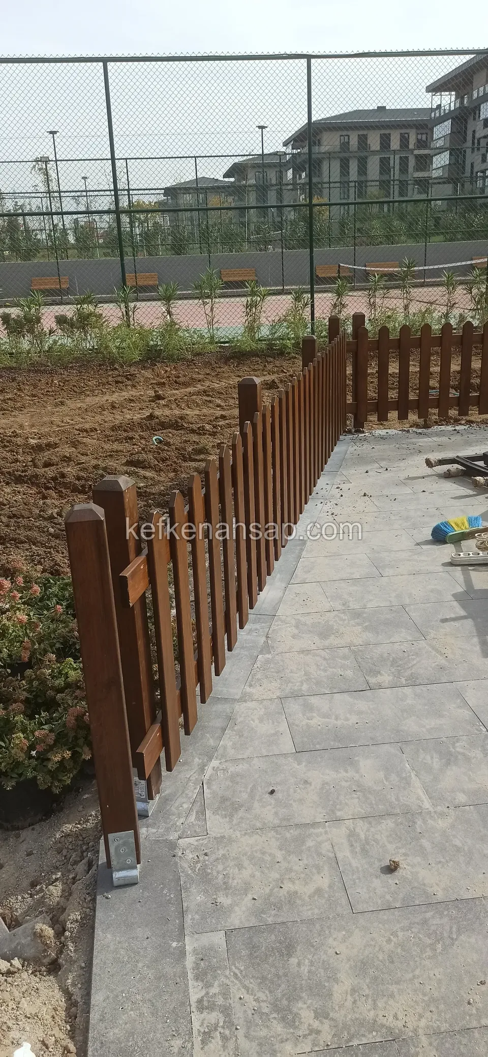 Gri taş zemin boyunca uzanan, metal ayakla sabitlenmiş koyu kahverengi çıtalı çit, arkada tel örgülü saha