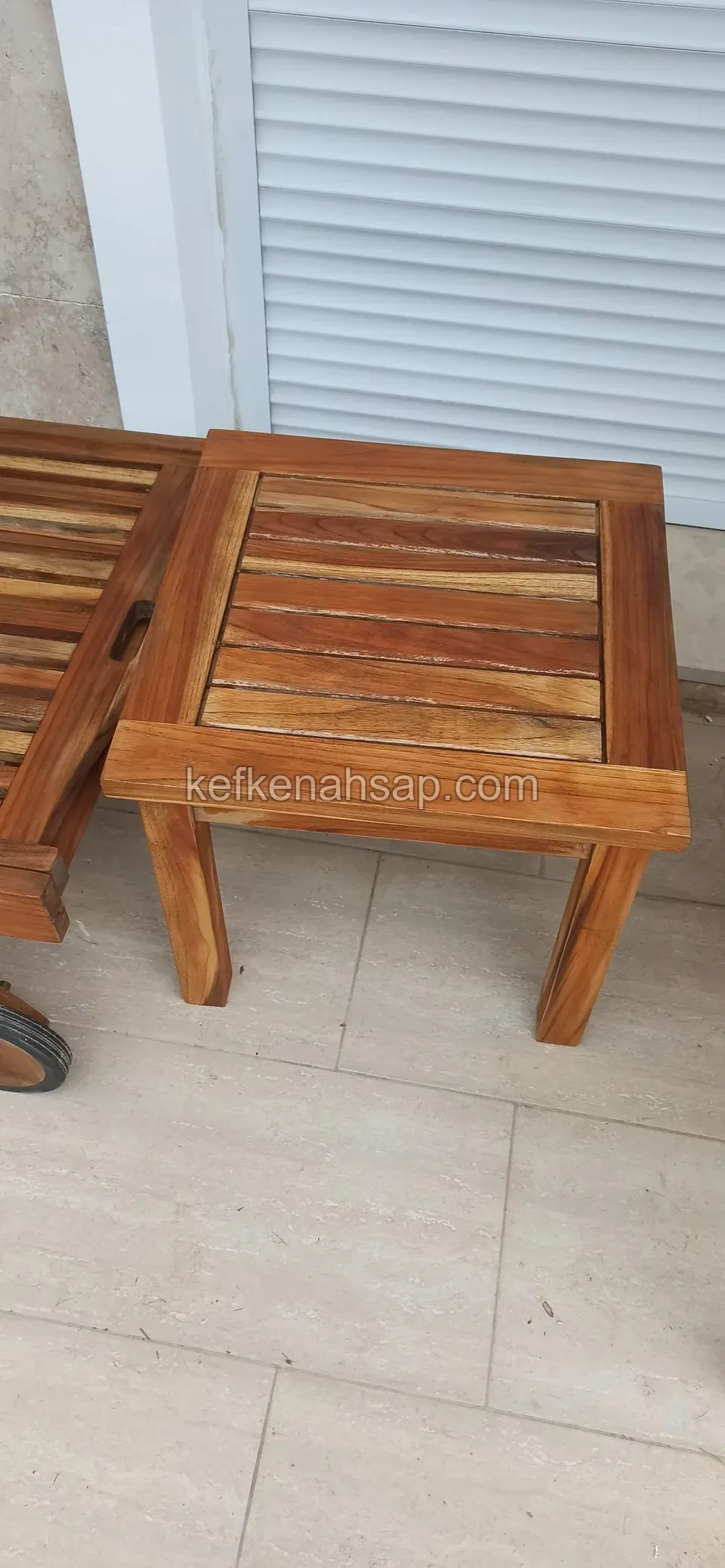 A small square timber table with a slatted framed top on tiled flooring in front of a wall with a closed roller shutter