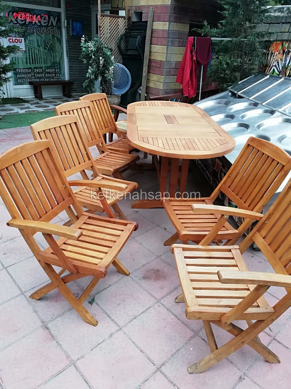 Auf Steinpflaster stehen ein ovaler Lattenesstisch aus Holz und rundum aufgestellte Klappstühle mit Lattenlehne und Armlehnen