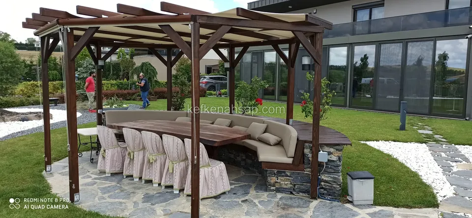 Taş döşeli yuvarlak zeminde tenteli ahşap pergola, altında kavisli minderli bank, uzun masa ve örtülü sandalyeler