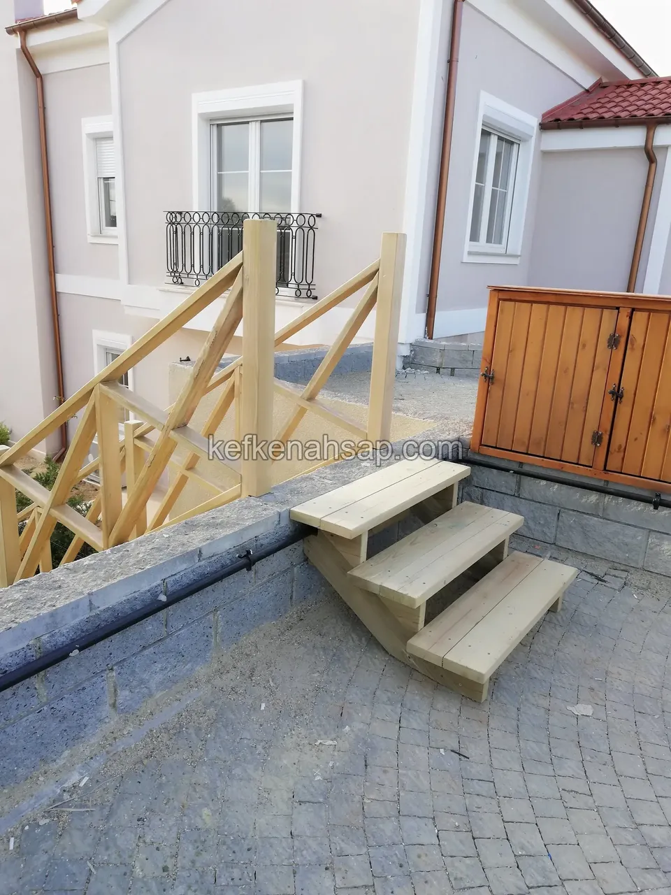Dreistufige Holztreppe auf Steinpflaster, dahinter ein Kreuzstreben-Geländer und ein Schrank mit Holztüren