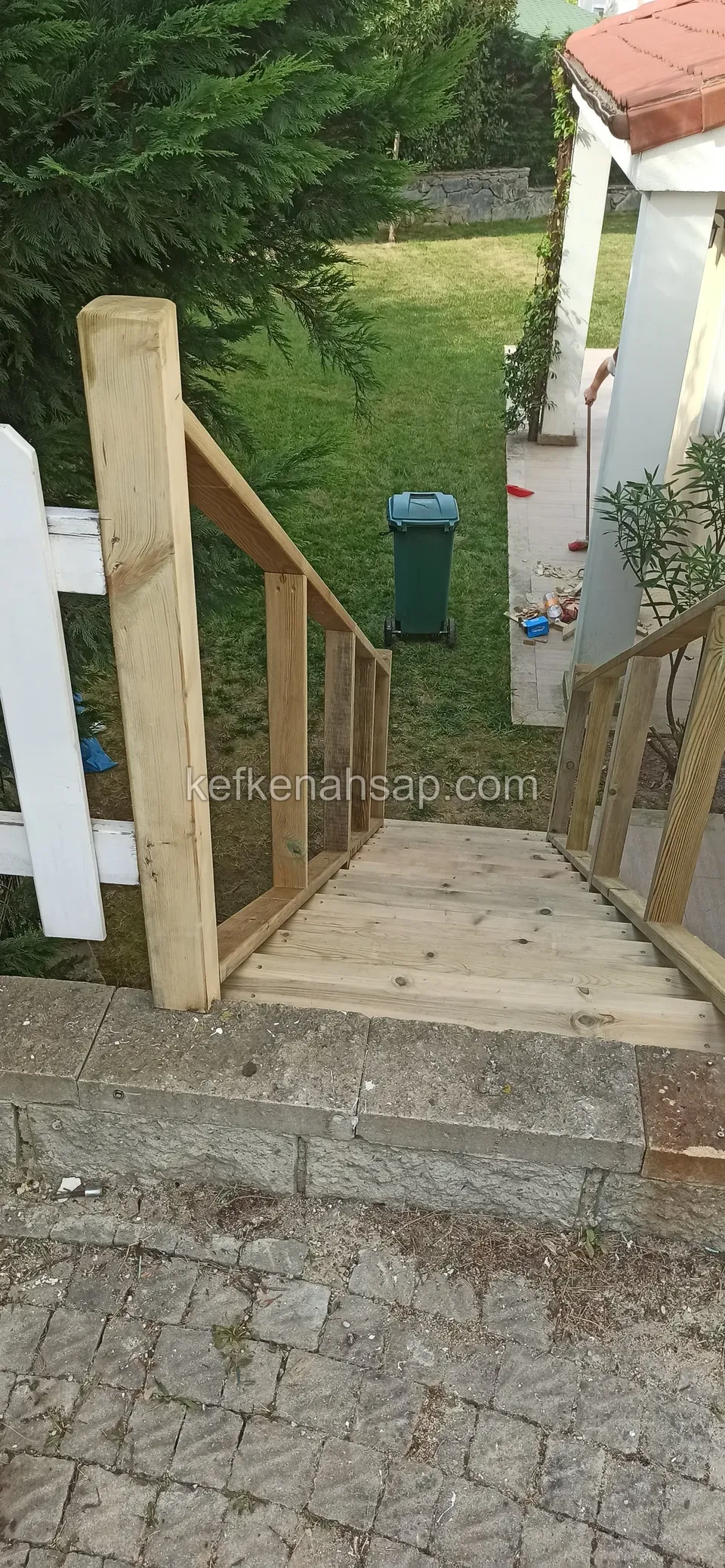 Neue Holztreppe mit Handlauf vom Steinpflaster hinab zum Rasen, links ein weißer Zaun