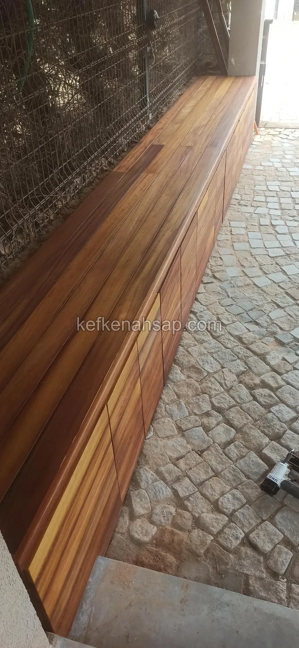 Tel çit önünde küp taş zeminde uzun ahşap kaplı dolap ve üst tablası