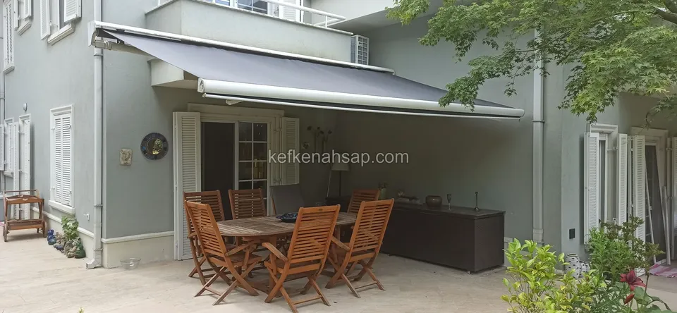 Auf der Terrasse eines Hauses steht unter einer Markise ein honigbrauner Holztisch mit Klappstühlen