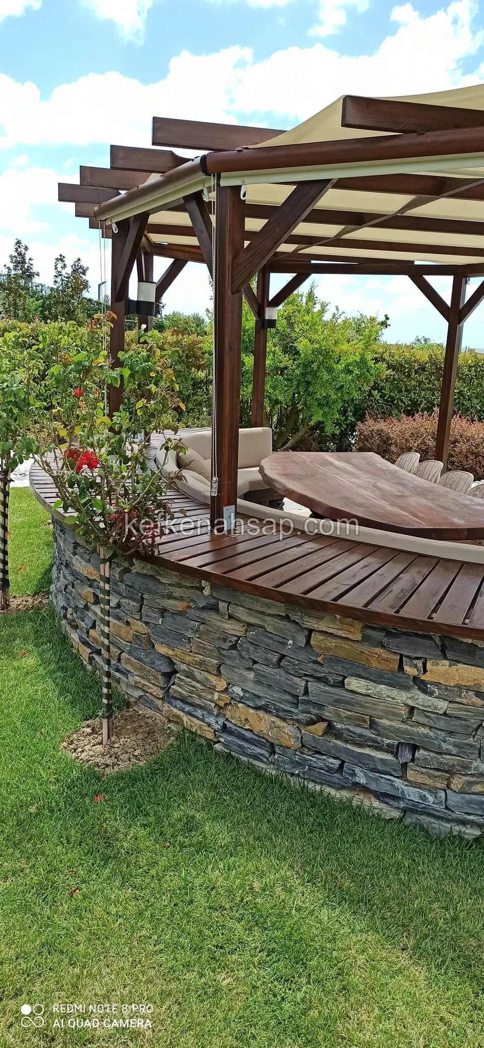 Gri-bej tenteli ahşap pergola, taş duvarlı kavisli bank, oval ahşap masa ve arkada çiçekli sarmaşıklar
