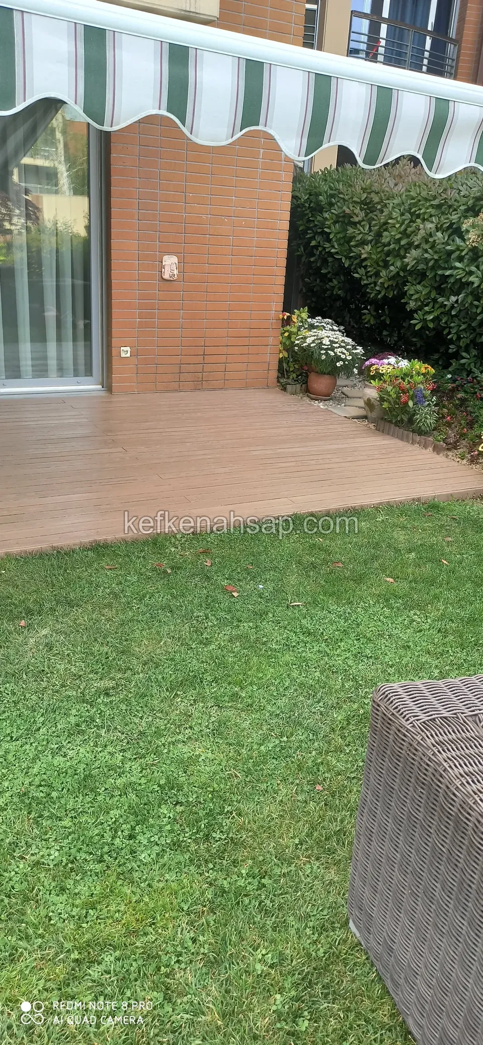 Light brown deck before the awning-covered entrance of a brick building, facing the lawn, wicker armchair at the edge