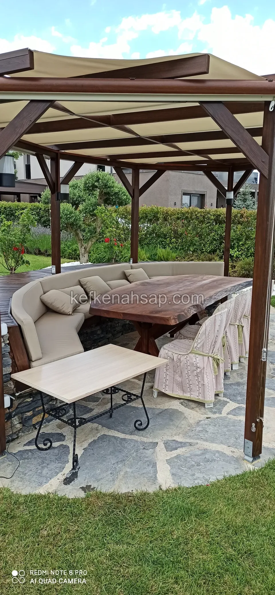 Tenteli pergola altında kavisli minderli oturma, ahşap masa ve ayakları metal bir sehpa, taş zemin