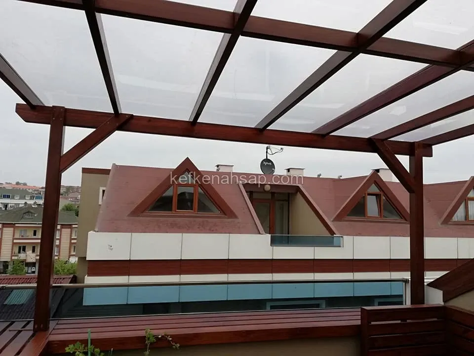 Teras üzerinde koyu kızıl vernikli ahşap pergola, kirişlerin üstünde şeffaf çatı paneli ve arkada çatı katlı evler