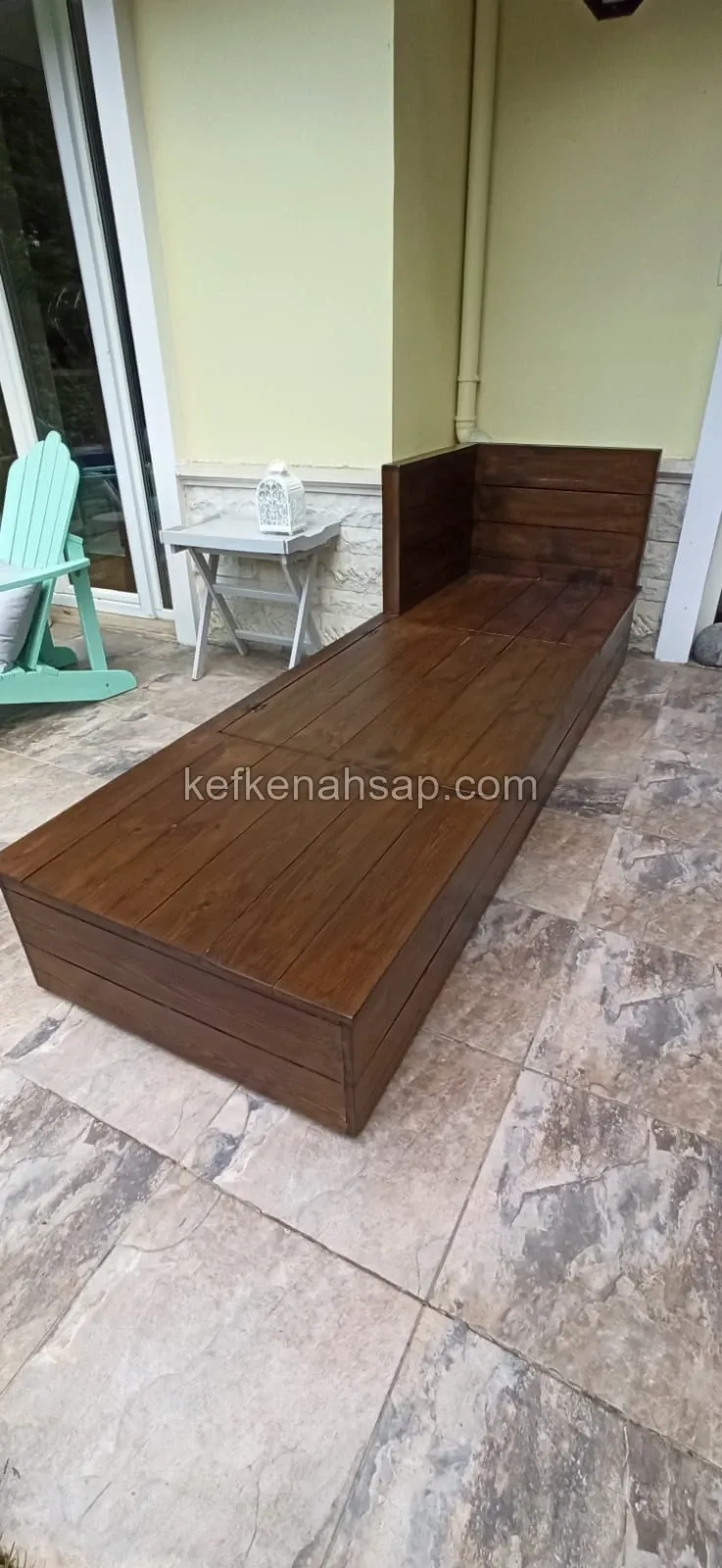 Long dark brown wooden bench with a raised back panel at one end, on a stone-tiled terrace