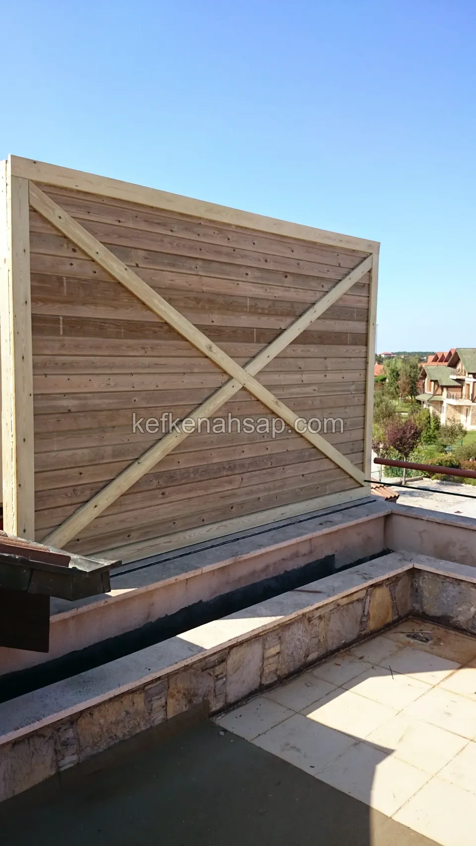 Çatı terasında taş bordür üzerinde X çaprazlı, yatay tahta kaplı yüksek ahşap perde, arkada yerleşim ve mavi gökyüzü