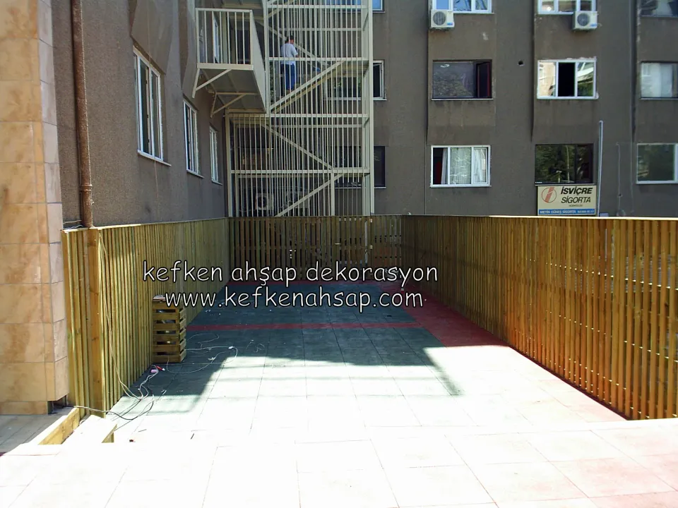 Apartman arkasındaki taş kaplı teras alanının iki yanında uzanan dikey çıtalı, açık tonlu ahşap çit
