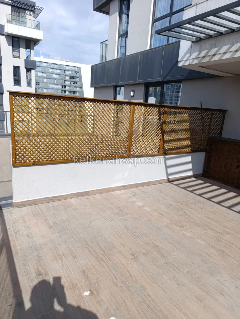 Apartman terasında beyaz duvar üzerine yerleştirilmiş, ceviz tonunda ahşap çerçeveli çapraz çıtalı panel ve sağda ahşap kapak