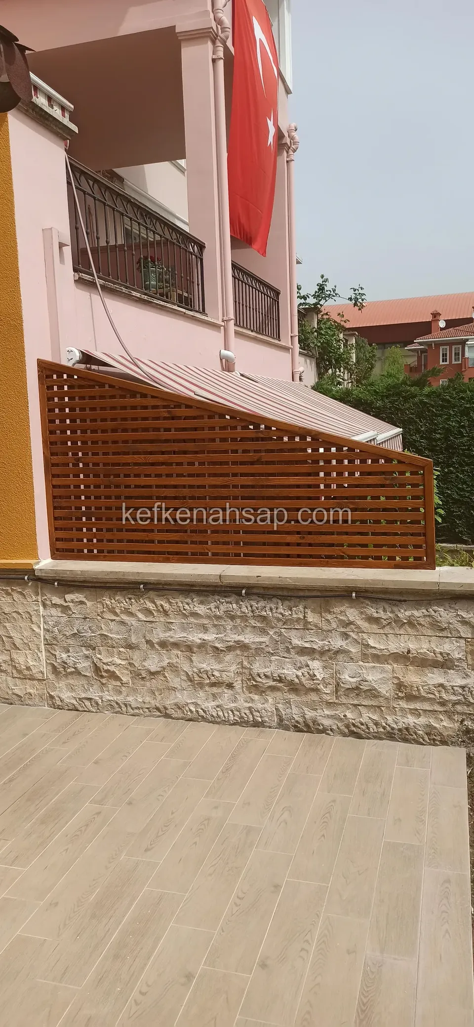Taş kaplı teras duvarının üstünde uzanan, sağa doğru alçalan yatay çıtalı turuncu ahşap panel