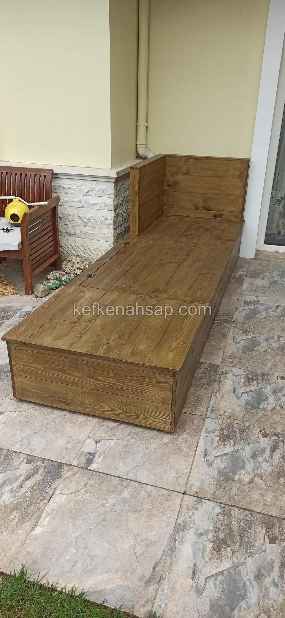 Eine lange, dunkel lackierte Holzliege mit hoher Lehne an einem Ende auf Terrassenfliesen in Steinoptik vor einer Hauswand