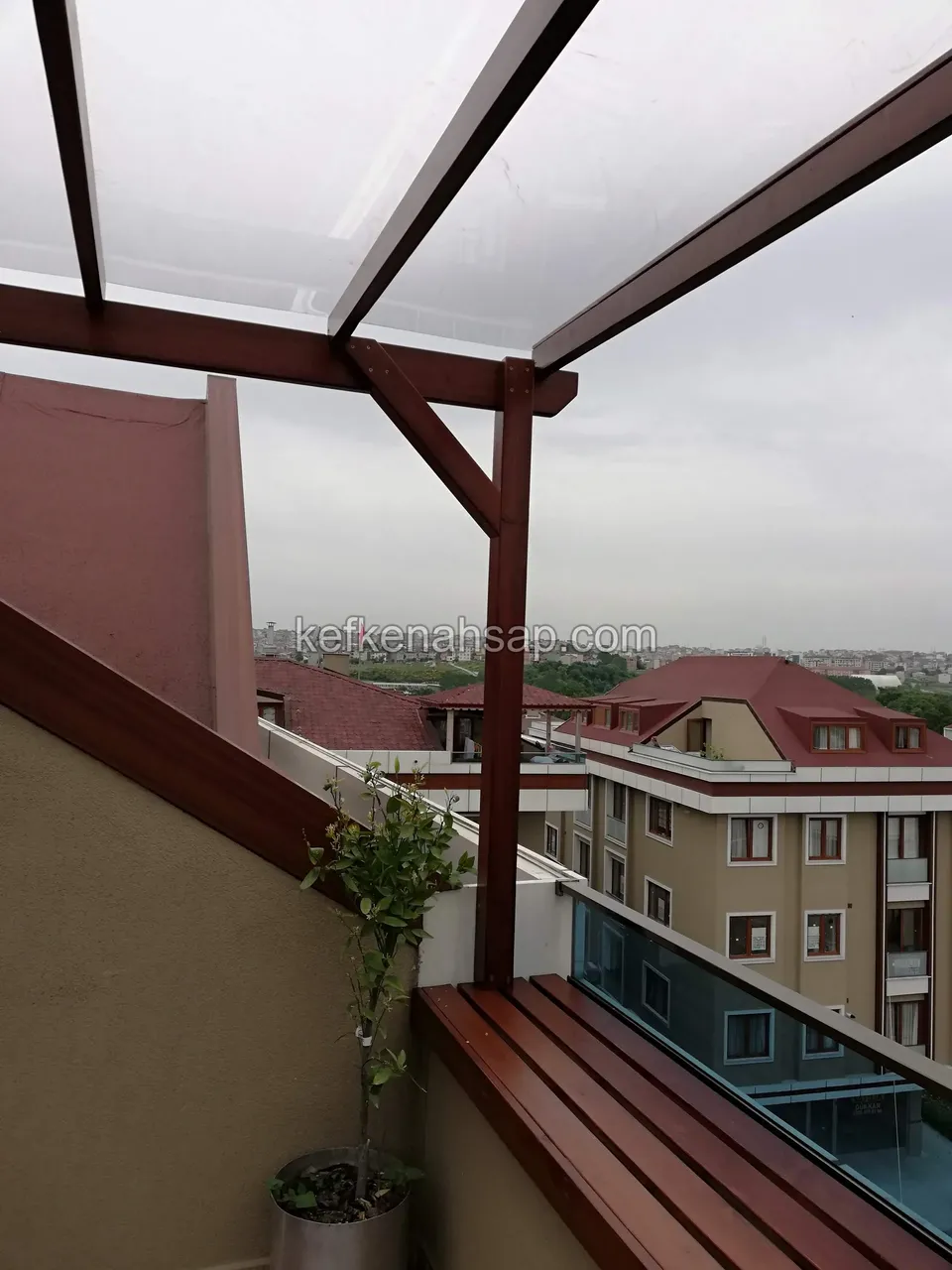 Teras köşesinde vernikli ahşap bank, kiriş ve direk üzerinde şeffaf çatı örtüsü, arkada apartmanlar