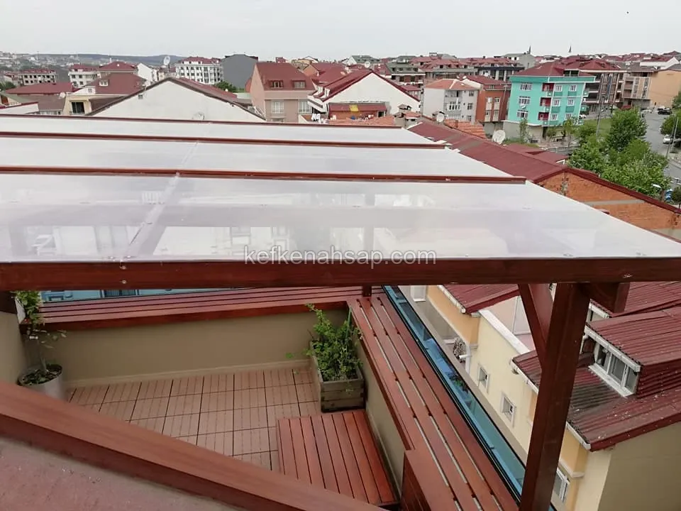 Çatı terasında koyu kahverengi ahşap taşıyıcılı pergola, şeffaf plaka örtü, ahşap oturma bankı ve saksılı döşeme