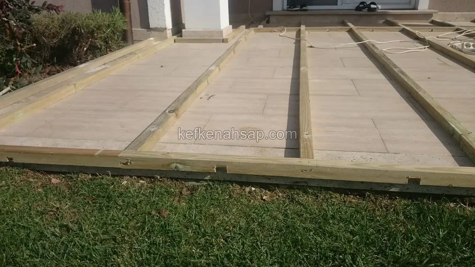 Evin girişindeki teras zemininde emprenyeli çam kirişlerle çerçevelenmiş geniş deck altlığı, önünde çimenlik