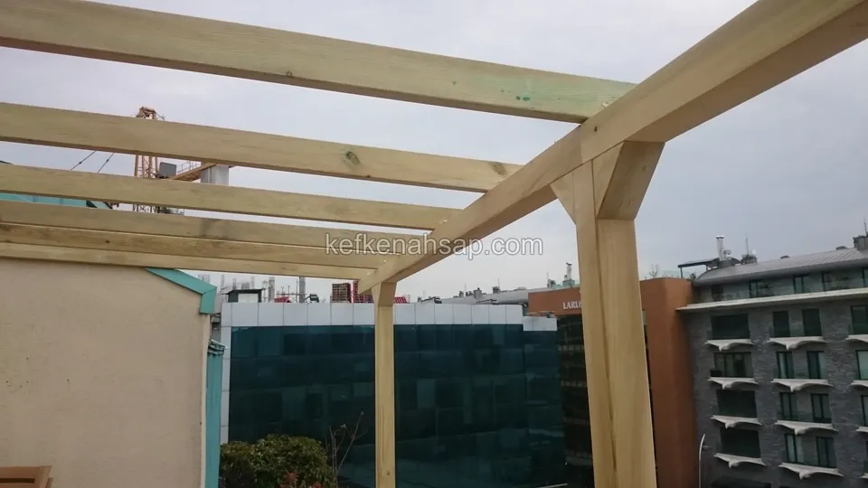 Terasın üzerinde emprenyeli direkli pergola iskeleti, payandalar ve arkada cam cepheli bina