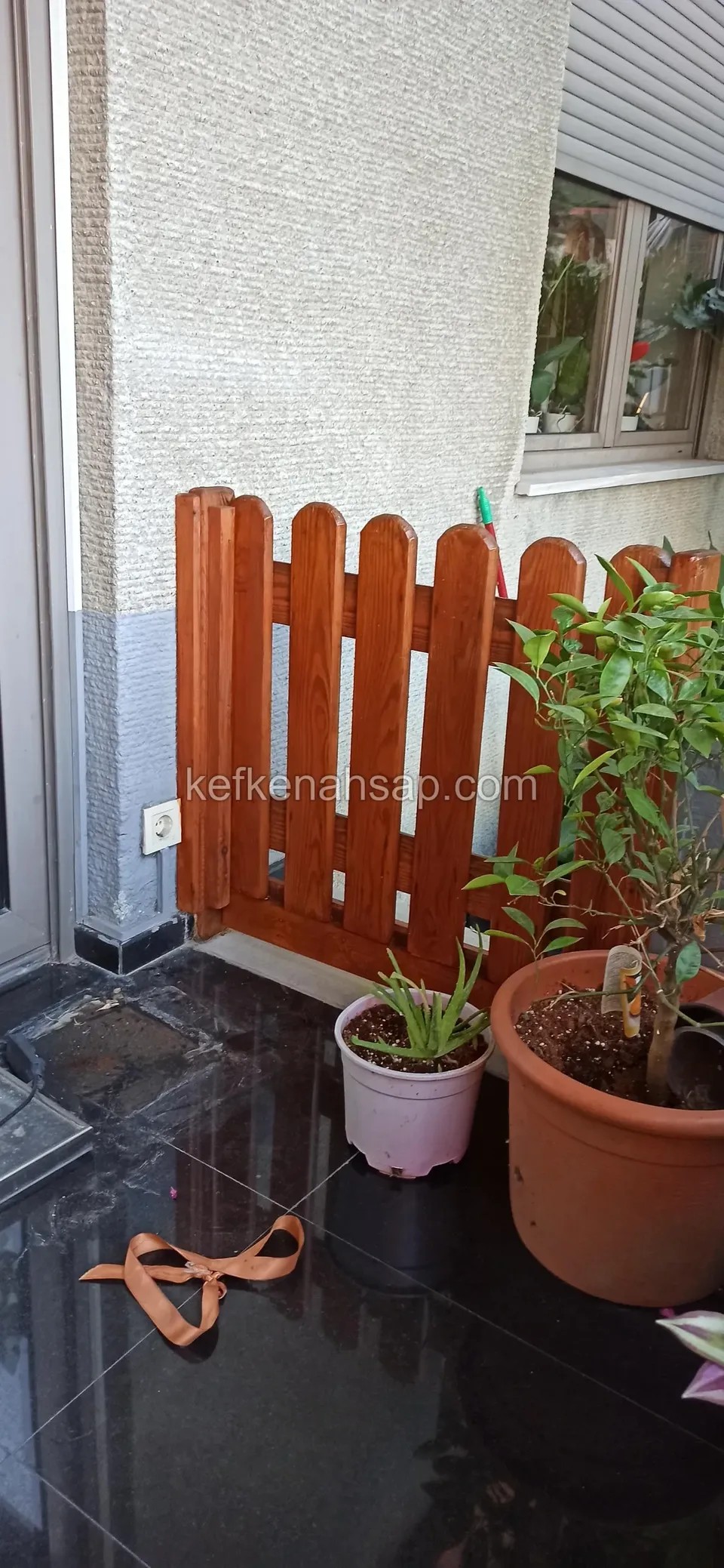 A low red-brown varnished pointed-slat timber fence beside potted plants in a corner of a black-tiled terrace