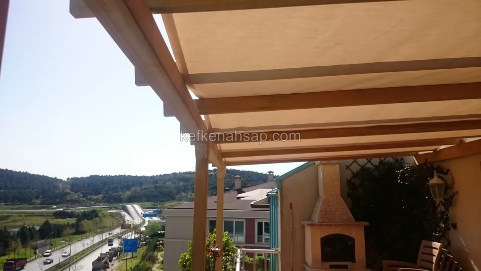 Terasta kumaş örtülü ahşap pergola, köşede tuğla şömine, ağaçlı tepeler ve aşağıda uzanan yol